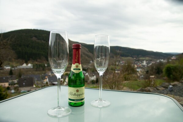 Una botella de vino Rottkäppchen y dos copas vacías sobre una mesa, con vistas a un pueblo y colinas al fondo.