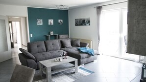 Un salon moderne avec un canapé d’angle gris, une table basse et une vue sur l’extérieur grâce à de grandes fenêtres.