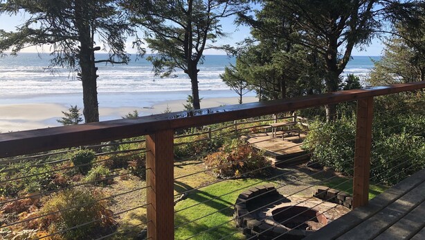 Eine Holzterrasse mit Blick auf das Meer und den Strand, umgeben von Bäumen und einer Rasenfläche mit einem Feuerkorb.