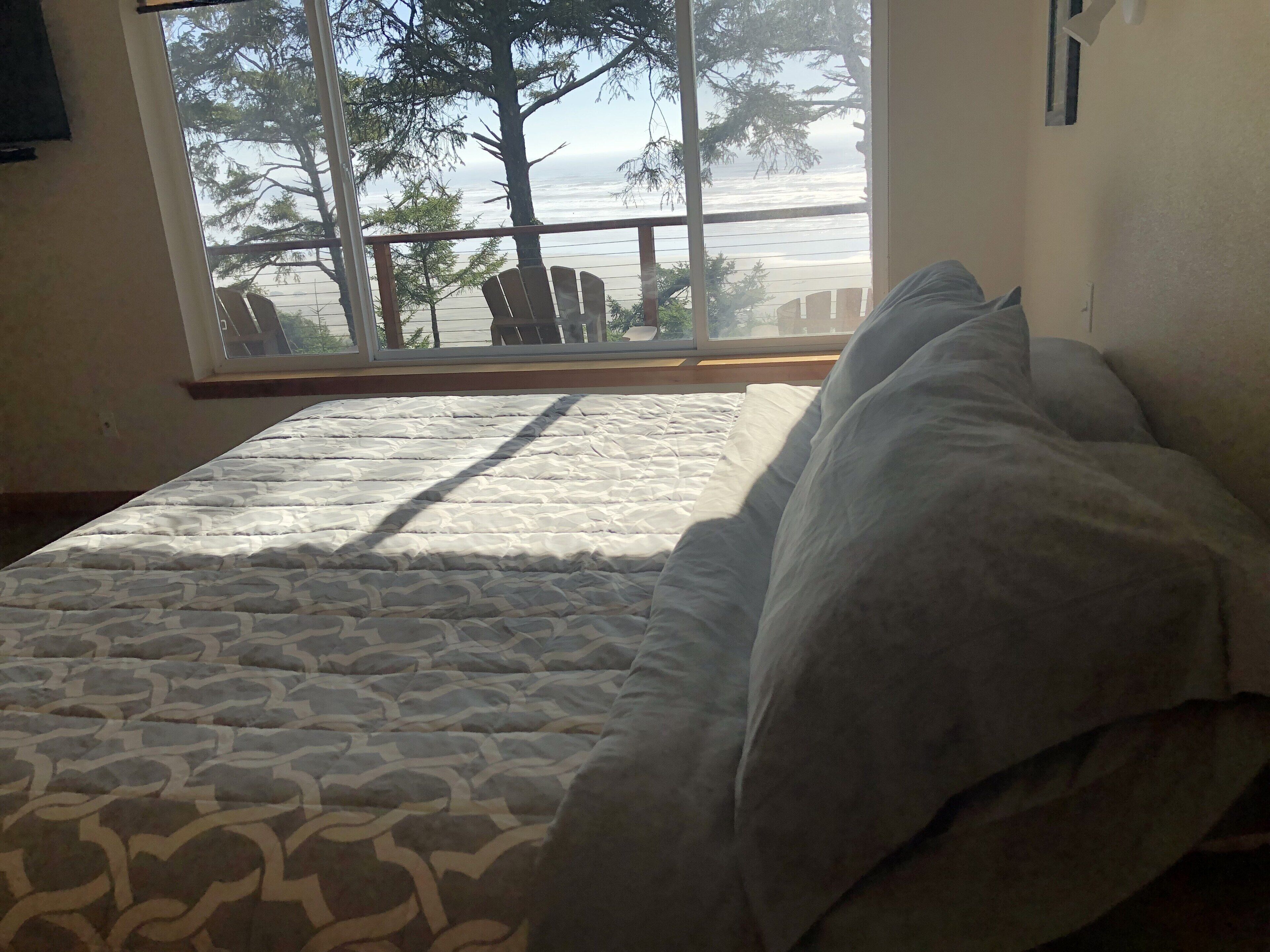 A bed with a patterned bedspread and pillows, a window with a view of the beach and trees, and a wooden headboard.