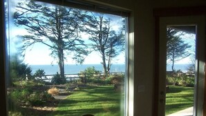 Ein Zimmer mit großem Fenster mit Blick auf einen grünen Rasen, Bäume und das Meer.