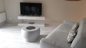 Ein modernes Wohnzimmer mit einem flachbildfernseher, der an einer weißen Konsole befestigt ist, einem runden Couchtisch und einem grauen Sofa mit einem gemusterten Kissen.