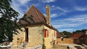 Un bâtiment en pierre, doté d’un toit en tuiles et de volets rouges, entouré d’un mur de pierre et d’une zone herbeuse.
