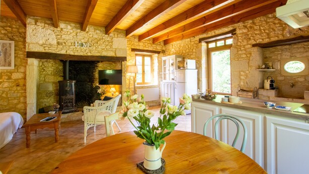 Un intérieur rustique avec des murs en pierre, des poutres en bois, une cheminée, un coin repas avec une table et des chaises, et une cuisine avec des meubles blancs et une fenêtre.