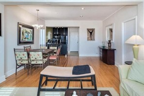 A living room with a sofa, a dining table with chairs, a kitchen in the background, and a lamp on a side table.