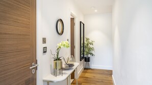 Un corridoio con una porta in legno, un muro bianco, uno specchio, una pianta in vaso e un vaso con fiori.