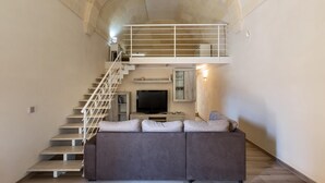 Ein großzügiger Wohnbereich mit hoher Decke, einer Treppe zu einer oberen Ebene, einem grauen Sofa, einem Flachbildfernseher und einem kleinen Schrank.