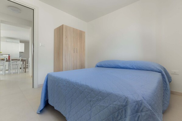A bedroom with a bed, a wooden wardrobe, and a kitchenette in the background.