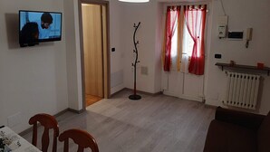 En stue med fjernsyn, spisebord med stole, sofa, radiator og et vindue med gardiner.
