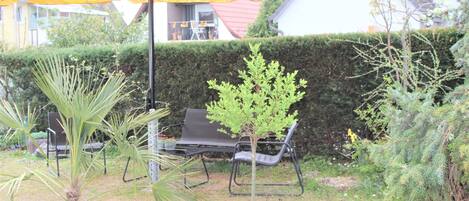 An outdoor seating area with a yellow umbrella, a small tree, and a hedge in the background.