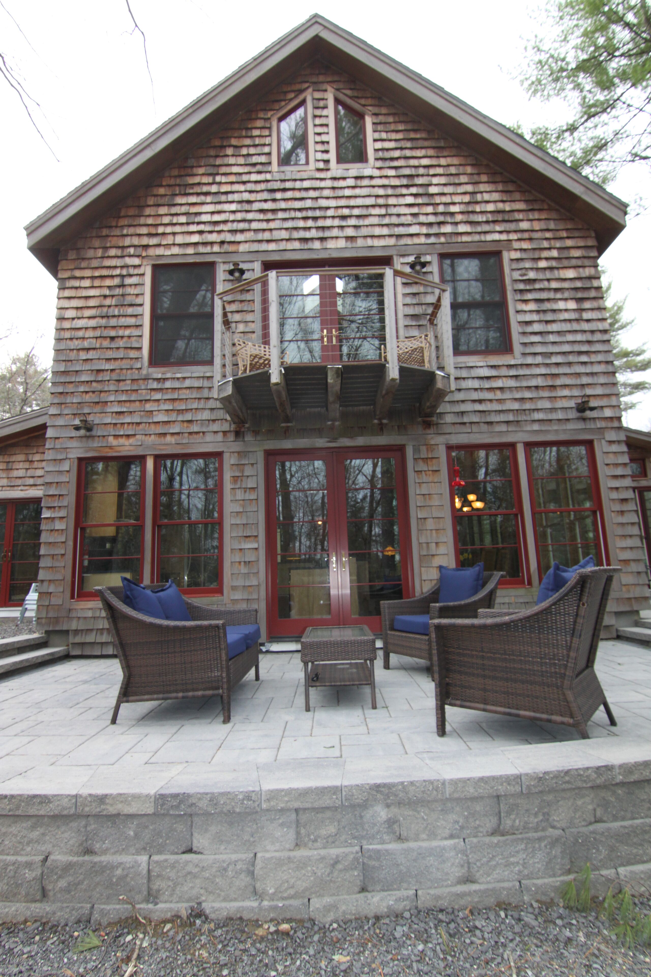 Une maison en bois à deux étages, dotée d’un balcon, de grandes fenêtres et de mobilier d’extérieur.