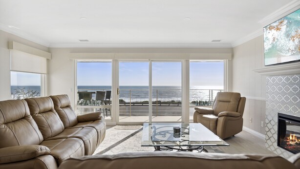 A spacious living room with a leather sofa, a glass coffee table, and a fireplace. Large windows offer a view of the ocean. - BEACH FACING! Large 3BR - 180 panoramic ocean views, steps to sand and Village! (Carlsbad)