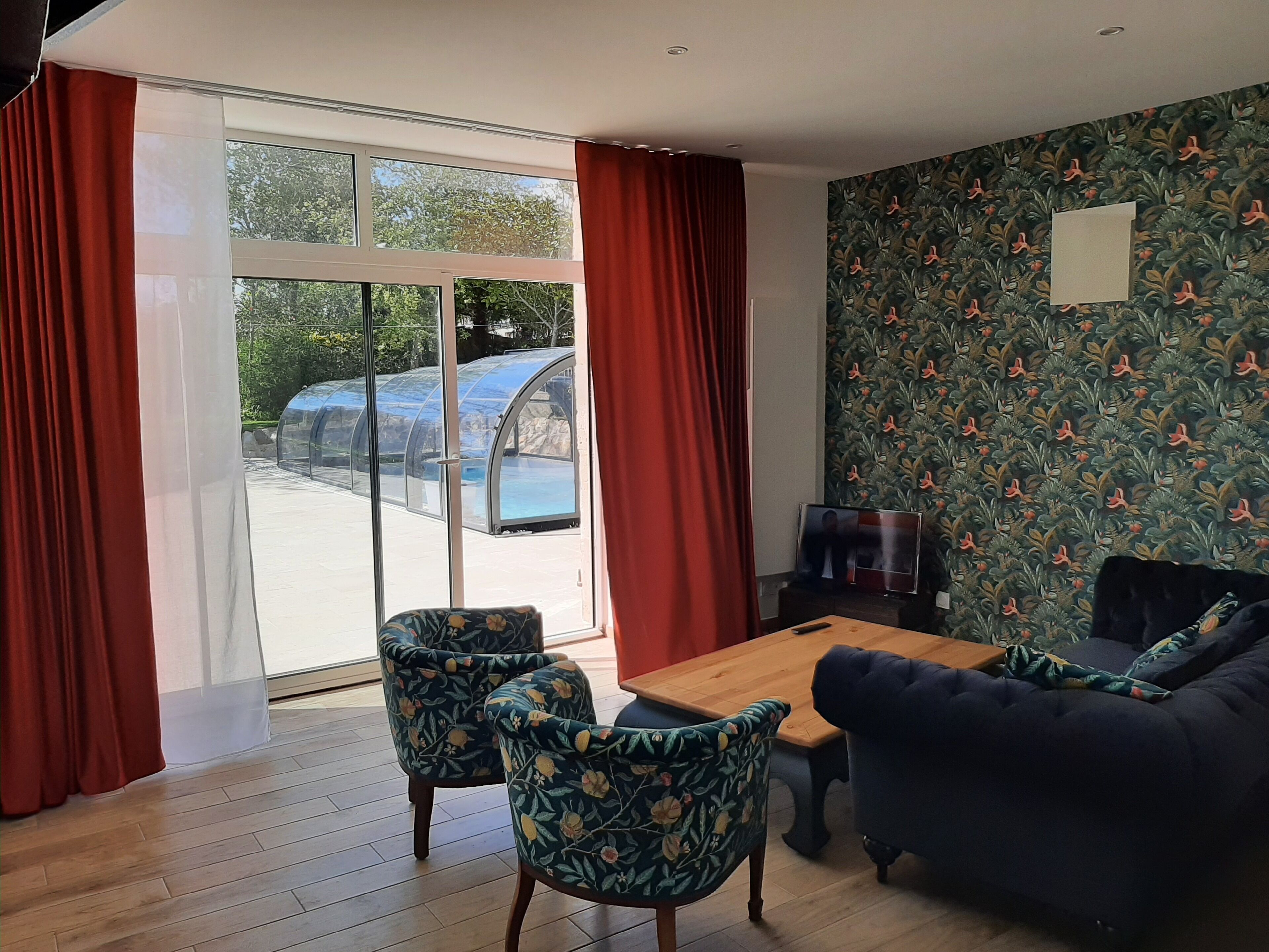 A room with a wooden floor, a dining table, two floral armchairs, a dark sofa, and a large window with red curtains.