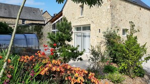 A stone building with a large window and a door, surrounded by a garden with various plants and flowers. - Fully renovated cottage with swimming pool in the heart of Cotentin (MONTEBOURG)