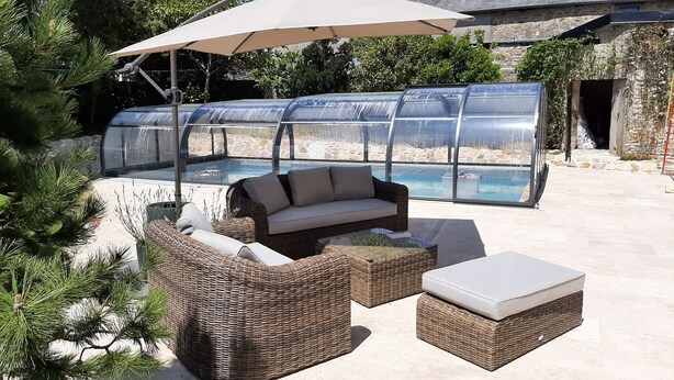 A patio area with a wicker sofa, ottoman, and umbrella, overlooking a pool with a glass enclosure. - Fully renovated cottage with swimming pool in the heart of Cotentin (MONTEBOURG)