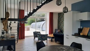 A modern kitchen with white cabinets, a dining table, and a black countertop. There is a staircase with black railings leading to a balcony. - Fully renovated cottage with swimming pool in the heart of Cotentin (MONTEBOURG)