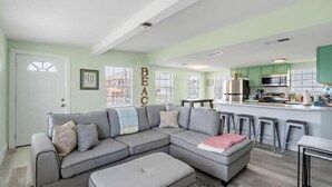 A living room with a grey sectional sofa, a small ottoman, and a kitchen area with green cabinets and a bar stool. - Cozy Cottage with Game Room, Close to Beach (Galveston)