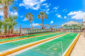 A craps table with a green felt surface, surrounded by a low concrete barrier, palm trees, and outdoor seating with umbrellas.