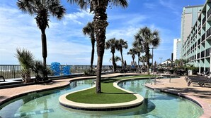A pool area with palm trees, lounge chairs, and a building in the background. - BEAUTIFUL PRIVATE 9TH FLOOR OCEAN VIEW CONDO; NEWLY RENOVATED OCEANFRONT RESORT (Myrtle Beach)