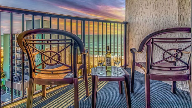 A balcony with two chairs and a table, a bottle of liquor, and a view of the cityscape. - BEAUTIFUL PRIVATE 9TH FLOOR OCEAN VIEW CONDO; NEWLY RENOVATED OCEANFRONT RESORT (Myrtle Beach)