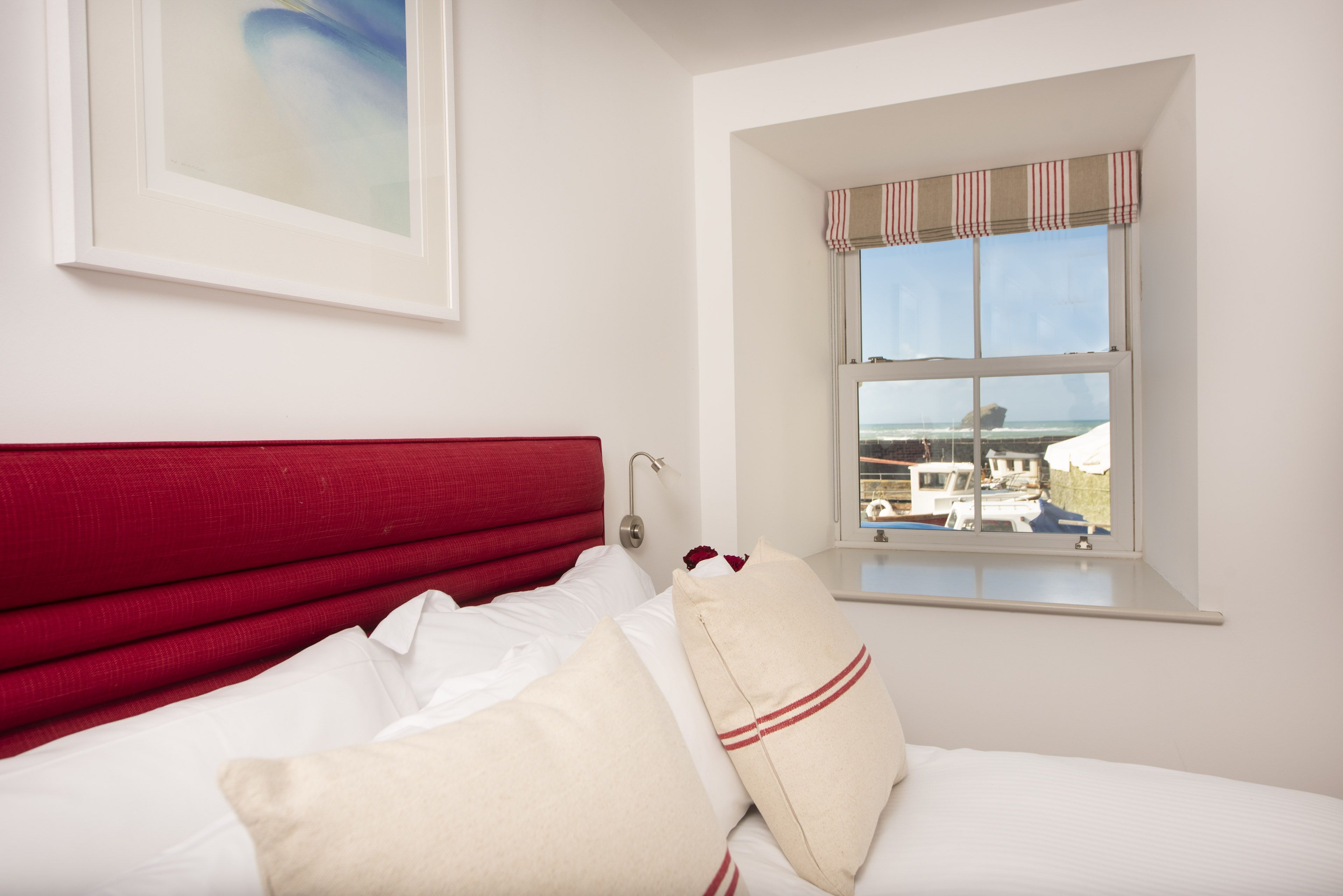 A hotel room with a red headboard, white bedding, a window with a view of boats, and a framed picture on the wall.