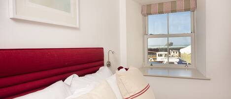 A hotel room with a red headboard, white bedding, a window with a view of boats, and a framed picture on the wall.