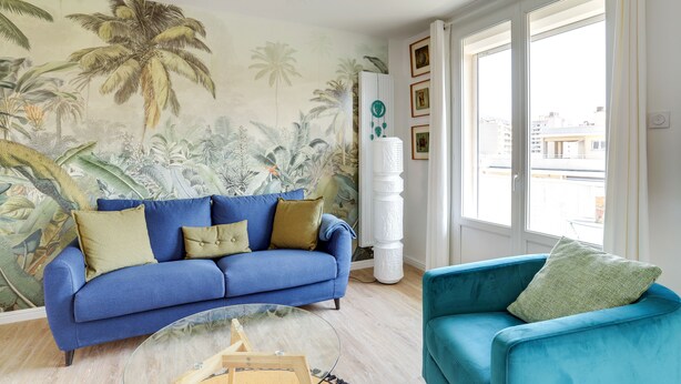 Ein Wohnzimmer mit einem blauen Sofa, einem türkisfarbenen Sessel und einem Wandgemälde mit tropischen Pflanzen.