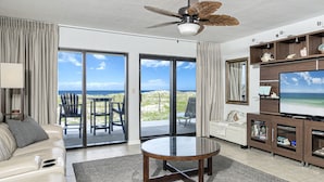 Un salon moderne avec un canapé, une table basse, un téléviseur et un ventilateur de plafond. De grandes fenêtres offrent une vue sur la plage et l’océan.