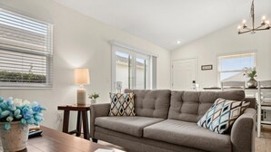 Ein Wohnzimmer mit grauem Sofa, einem Holztisch und einer Lampe. Es gibt ein Fenster mit Jalousien, eine Tür und einen Kronleuchter.