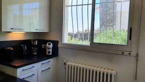 A kitchen with white cabinets, a black countertop, a coffee maker, a kettle, and a window with a view of greenery and buildings. - Villa Tanchet-Plage Sea view (Les Sables-d'Olonne)