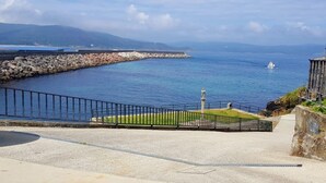 A coastal view with a paved walkway, a stone wall, and a grassy area with a metal fence. - Apartamento Playa da Ribeira Finisterre (Fisterra)