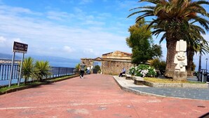 A paved walkway leading to a historic building with a stone facade, palm trees, and a sign indicating the location. - Apartamento Playa da Ribeira Finisterre (Fisterra)