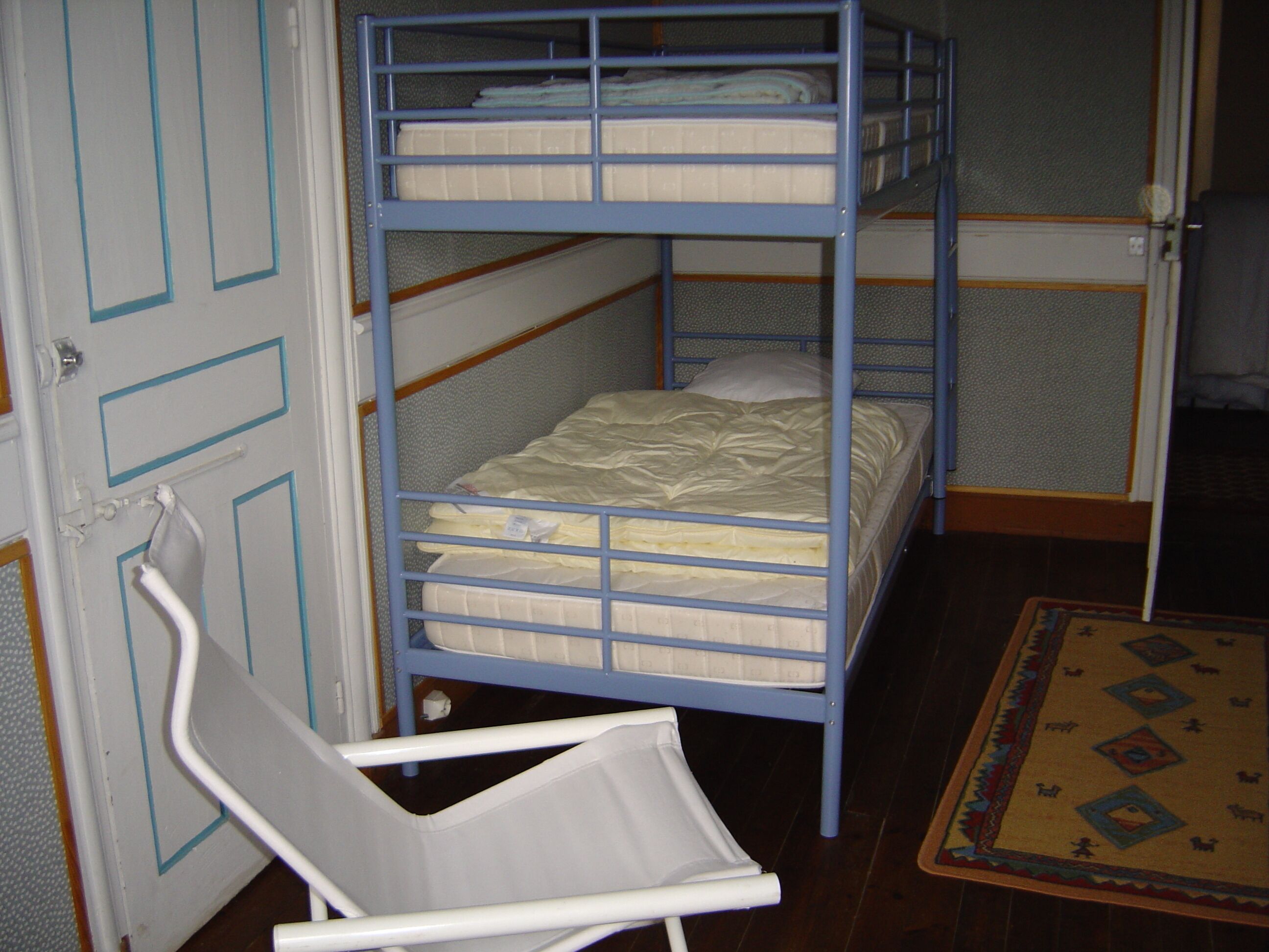 Una cama litera con colchón y almohadas, una silla blanca y una alfombra estampada en el suelo.