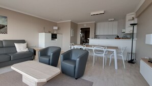 Un salon moderne avec un canapé gris, deux fauteuils, une table basse et un coin repas comprenant une table en bois et des chaises blanches.
