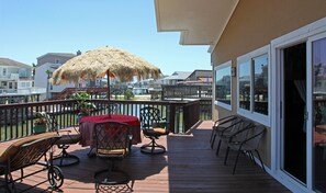 Eine Holzterrasse mit Tisch und Stühlen, einem Reetdachschirm und Blick auf Wasser und Gebäude im Hintergrund.