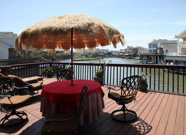 Eine Terrasse mit Tisch und Stühlen, einem reetgedeckten Sonnenschirm und Blick auf einen Fluss und Gebäude.