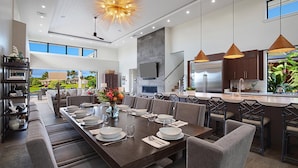 A modern dining area with a long table set for six, surrounded by chairs, a chandelier, and a view of the outdoors. - Spectacular home with ocean and golf course views! (Koloa)