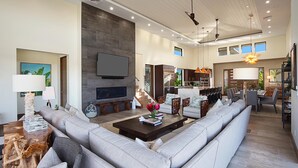 A spacious living room with a large sectional sofa, a coffee table, and a fireplace. There is a dining area with a kitchen in the background. - Spectacular home with ocean and golf course views! (Koloa)