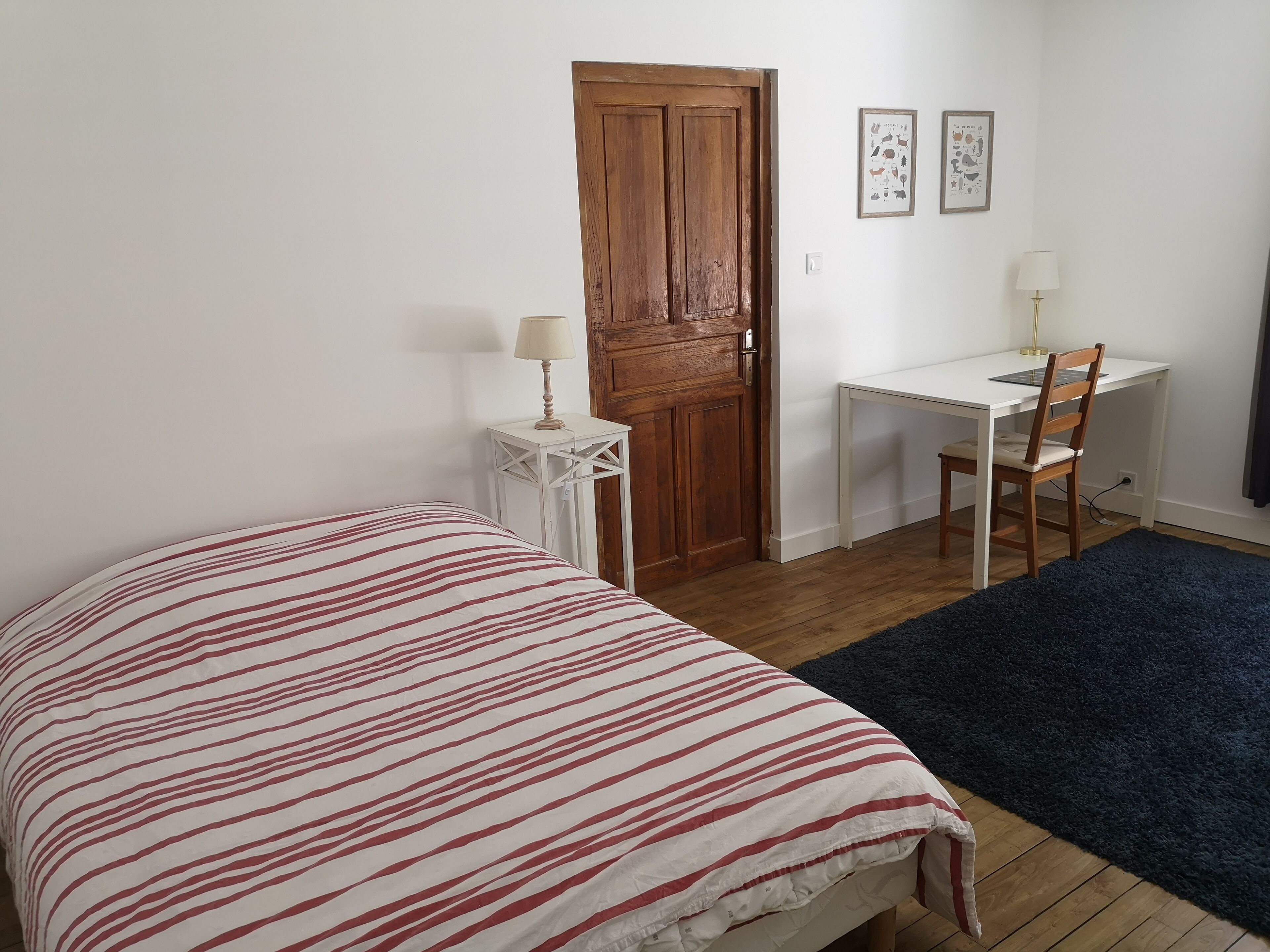 A bedroom with a bed, a desk, a chair, a wooden door, and a small white table with a lamp.