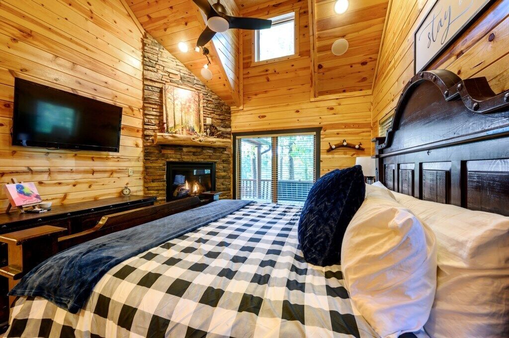 A cozy log cabin bedroom with a checkered bedspread, a stone fireplace, a flat-screen TV mounted on the wall, and a large window providing a view of the outdoors.