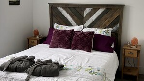 Una cama bien hecha con edredón blanco y varios almohadones, cabecera de madera y dos mesitas de noche con objetos decorativos.