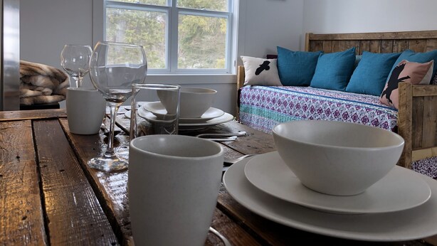 Una habitación acogedora con una mesa de madera preparada para comer, una cama con ropa de cama azul y estampada, y una ventana que da a árboles.