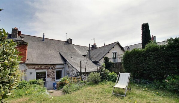 A stone house with a sloped roof, a garden with a chair, and a hedge.