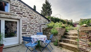 Edificio de piedra con un pequeño patio que incluye una mesa, sillas, una puerta y escalones que suben a un jardín.