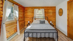 Une chambre située dans une cabane en rondins, avec un lit en métal, deux tables de chevet avec des lampes et une grande fenêtre avec des rideaux.
