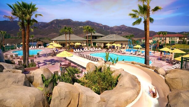 Un resort con una amplia zona de piscina, palmeras y montañas al fondo.