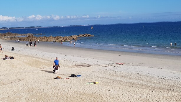 Una playa con gente caminando y nadando, un rompeolas rocoso y un cielo azul despejado.