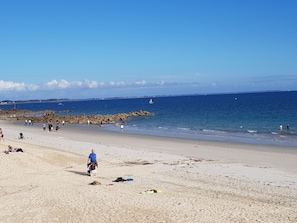 En strand med folk som promenerar och badar, en stenbrytande vågbrytare och en klarblå himmel.