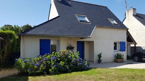 Una casa de dos pisos con techo oscuro, puertas azules y un jardín con flores moradas.