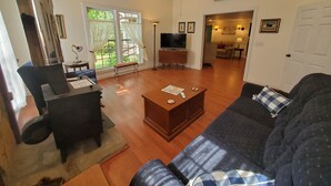 A living room with a dark blue sofa, wooden coffee table, and a TV stand. There is a ceiling fan, a large window with curtains, and a door leading to another room. - Mountain Laurel Cottage ~ Bat Cave North Carolina  RELAX, REFRESH, RENEW! (Hendersonville)